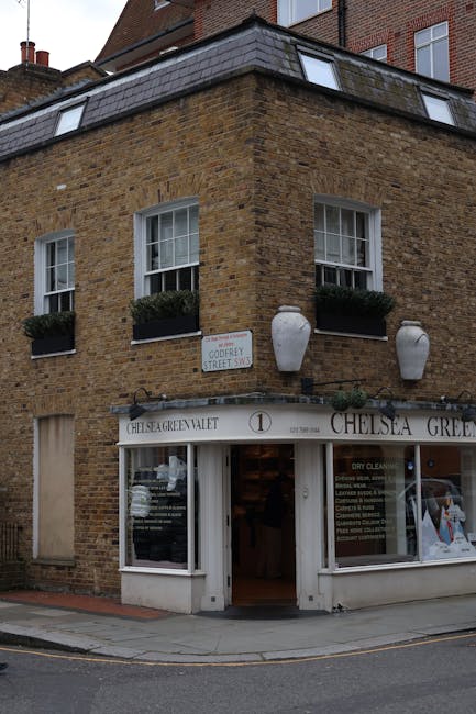 The image shows a corner building constructed with light brown brickwork, featuring a shop window on the ground floor displaying cleaning services and signage for Chelsea Green Valet. Above the shop, 